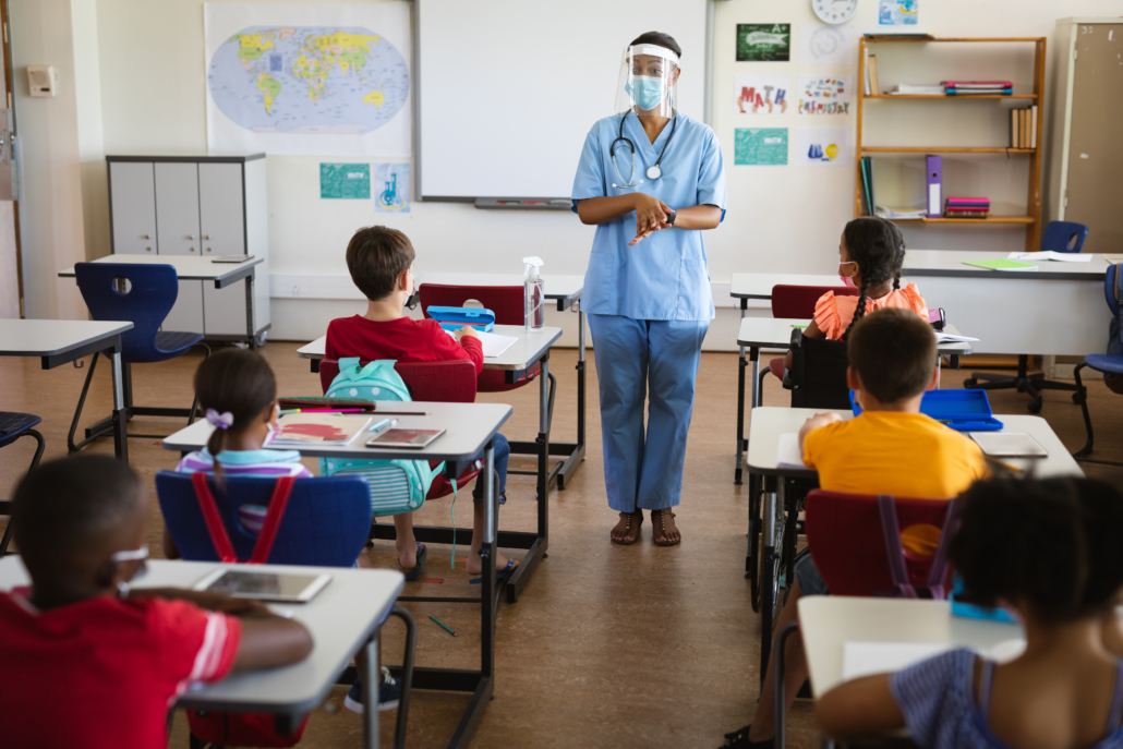 Une professionnelle de la santé portant un écran facial parlant à des élèves en classe dans une école primaire