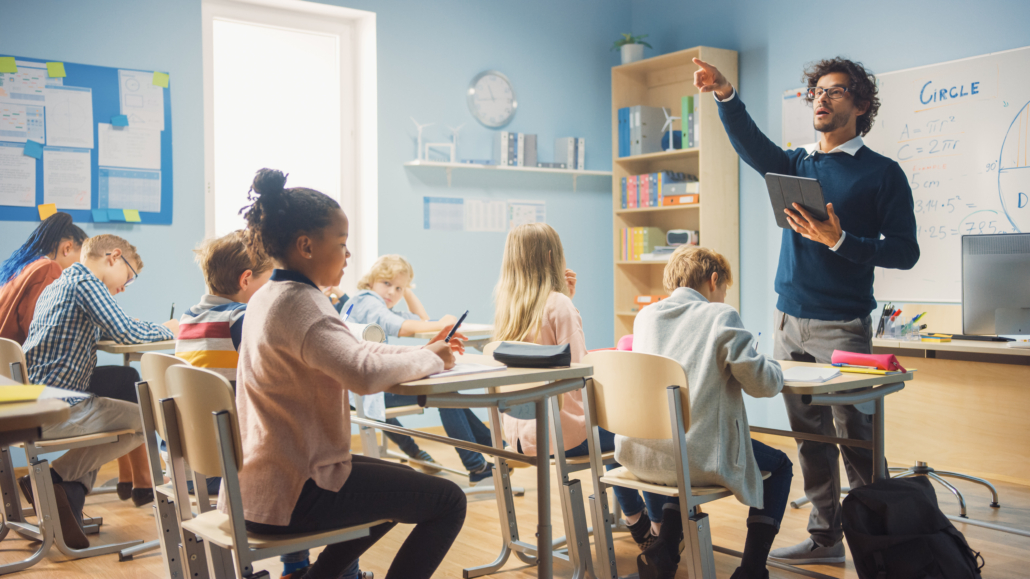 Enthusiastic Teacher Giving a Lesson in Elementary School to Class Full of of Diverse Children. Group of Smart Multiethnic Kids Learning New Stuff. Side View Shot